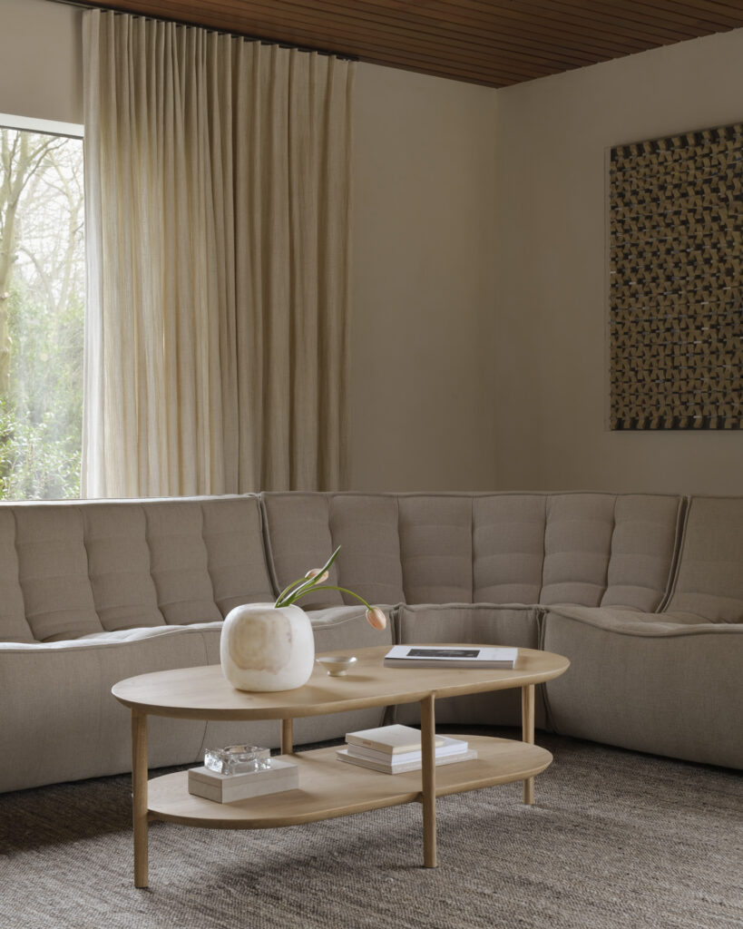 Un salon minimaliste comprend un canapé sectionnel beige, une table basse ronde en bois avec un vase blanc et un livre, des rideaux beiges, une tenture murale tissée et une grande fenêtre donnant sur la verdure. Adjacent au salon se trouve une élégante table à manger carrée Bok qui complète le décor serein.