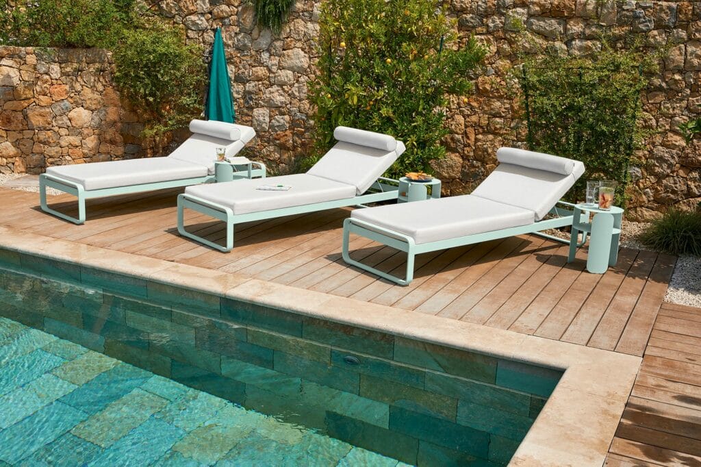 Trois transats blancs au bord de la piscine et une table d'appoint Bebop sur une terrasse en bois à côté d'une piscine avec un fond de mur en pierre et de verdure.