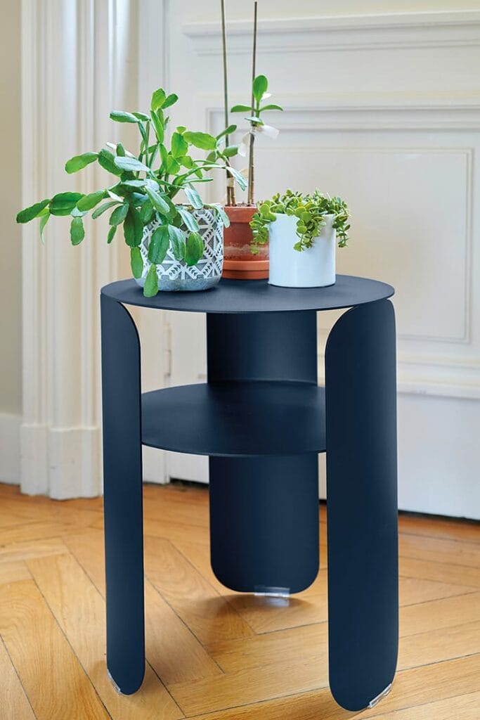 Une table d'appoint Bebop noire moderne avec des pieds courbés, contenant trois plantes en pot, positionnée sur un plancher en bois contre un mur blanc.