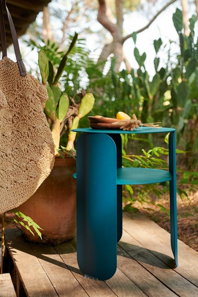 Une Table d'appoint Bebop en métal bleu sur laquelle est accroché un sac de paille, posée sur une terrasse en bois entourée d'une verdure luxuriante.