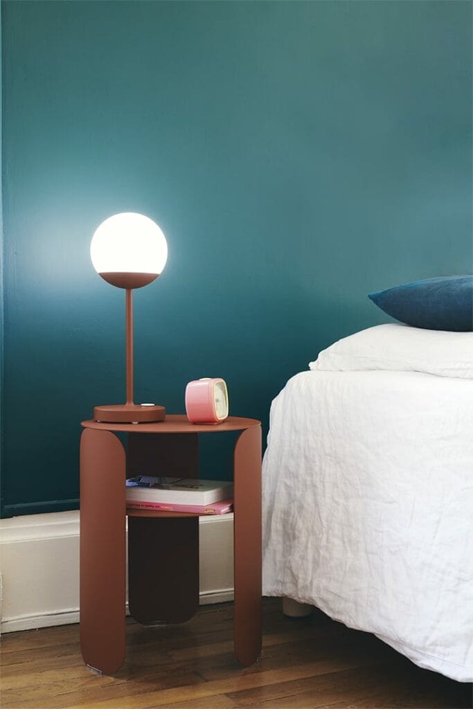 Une table de chevet moderne avec une lampe ronde blanche sur une table d'appoint Bebop à côté d'un lit, avec des livres et un objet rose sur la table contre un mur vert foncé.