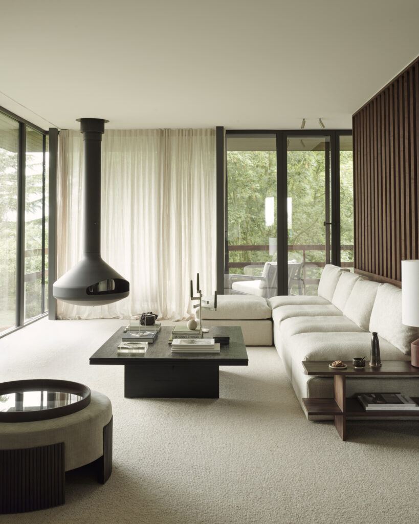 Salon moderne avec de grandes fenêtres, des meubles de couleur claire, une cheminée suspendue noire, un canapé sectionnel, une table basse et une décoration minimaliste. L'espace est bien éclairé par la lumière naturelle et dispose de l'élégant Pouf Roller Max pour des sièges supplémentaires ou pour se détendre.