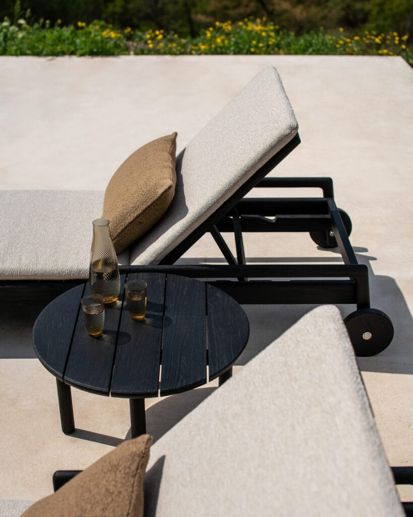 Une chaise longue en teck teinté noir et une table d'appoint de jardin sur un patio.