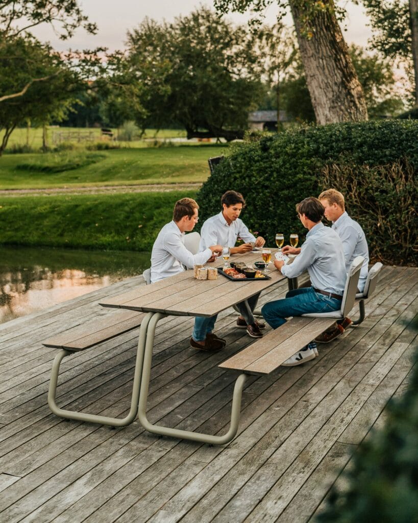 Quatre personnes profitant d'un repas en plein air près d'une table gris clair au bord d'un étang au crépuscule.