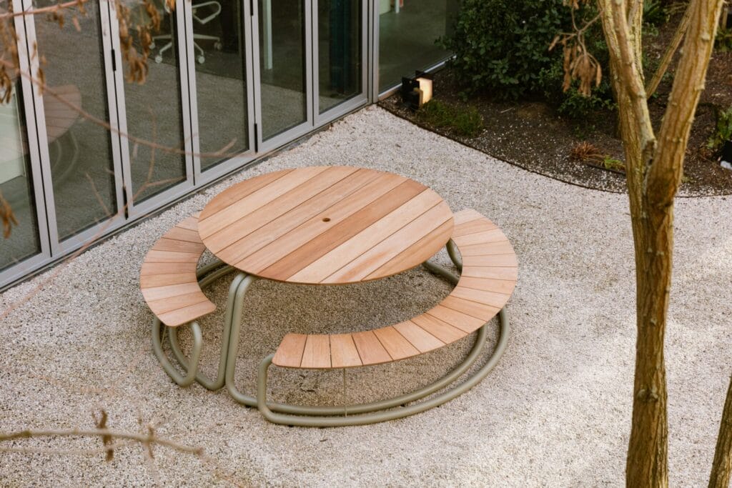 The Circle - table d'extérieur en bois avec banquettes incurvées intégrées sur une surface de gravier gris clair près d'un bâtiment avec des portes en verre.