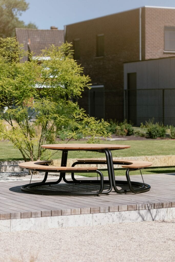 Coin salon extérieur avec un banc incurvé moderne et une table The Circle sur une plate-forme en bois entourée de verdure, avec des accents gris clair RAL7044.