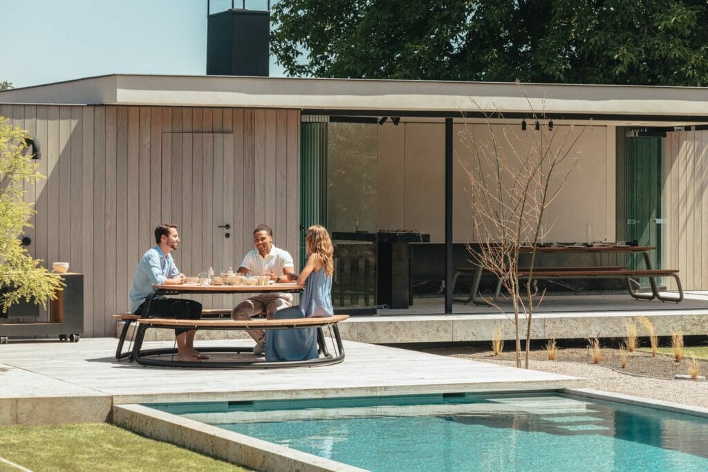Trois personnes prenant un repas au Circle ont dressé une table avec des bancs en gris clair (RAL7044) au bord d'une piscine, avec une maison moderne en arrière-plan.