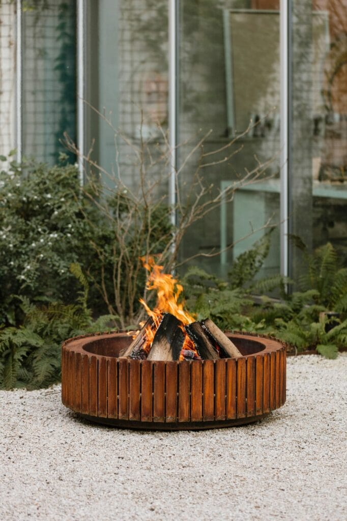 Un feu brûlant dans un foyer rond en acier Corten rouillé, avec un fond de verdure luxuriante et un bâtiment en verre moderne.
