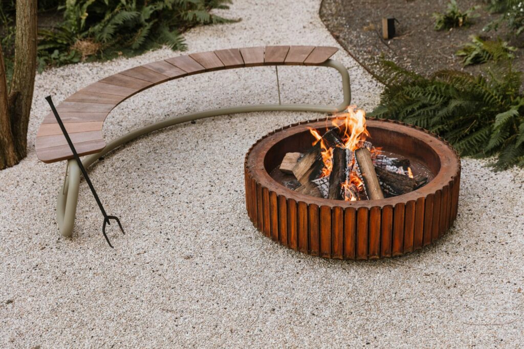 Un banc en bois courbé moderne entourant un foyer en acier Corten dans un décor extérieur en gravier.