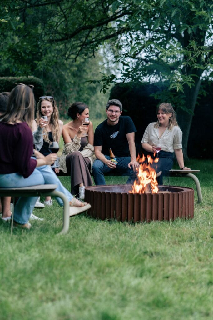 Groupe d'amis prenant un verre et riant autour d'un foyer en acier Corten dans un jardin.