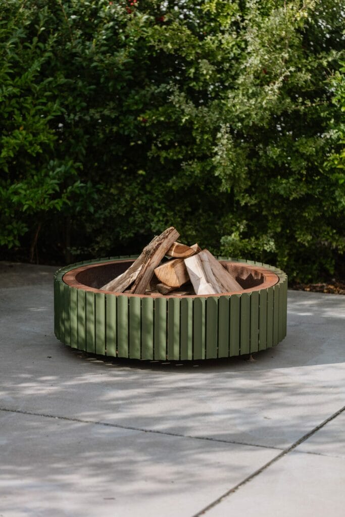 Un foyer rond en acier Corten contenant des bûches, posé sur une surface en béton avec de la verdure Vert Olive en arrière-plan.