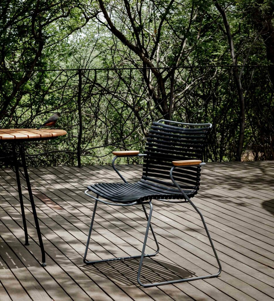 Une table, des chaises et une chaise Click avec accoudoirs sur une terrasse en bois dans un espace boisé.