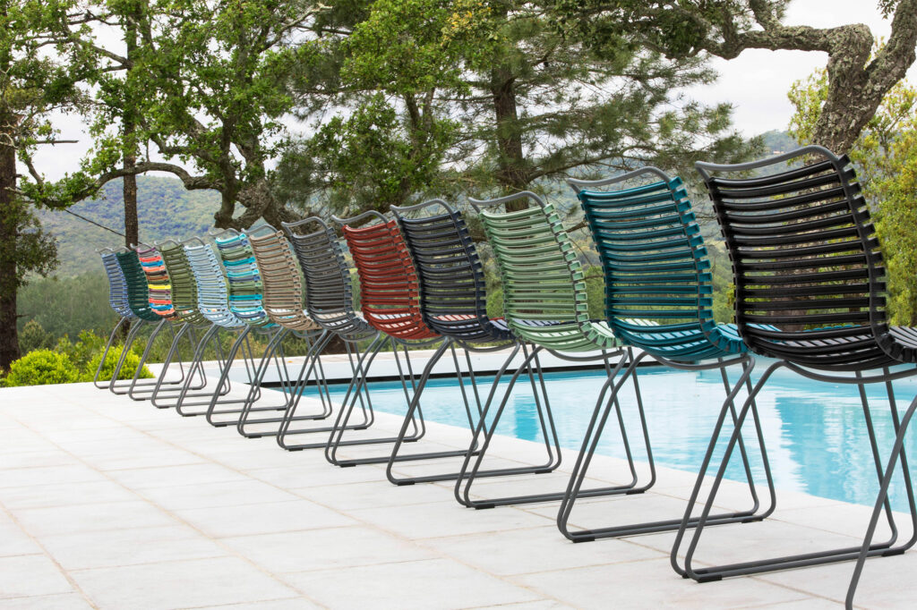 Une rangée de chaises CLICK Dining colorées sans accoudoirs alignées au bord d’une piscine, avec en toile de fond des arbres et des collines vert pin.