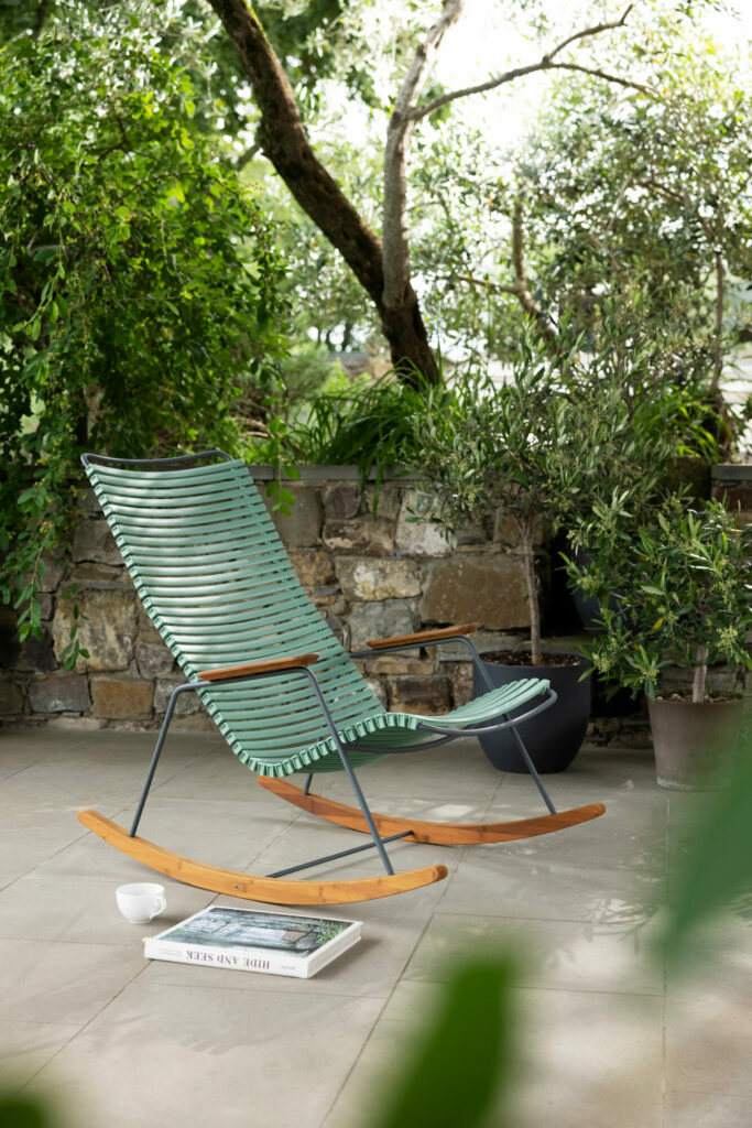 Un fauteuil à bascule vert Click dans un jardin tranquille avec des livres posés au sol à proximité.