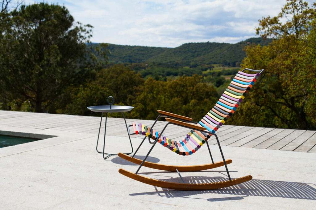 Un fauteuil à bascule Click avec une petite table ronde sur une terrasse au bord de la piscine avec un paysage verdoyant en arrière-plan.