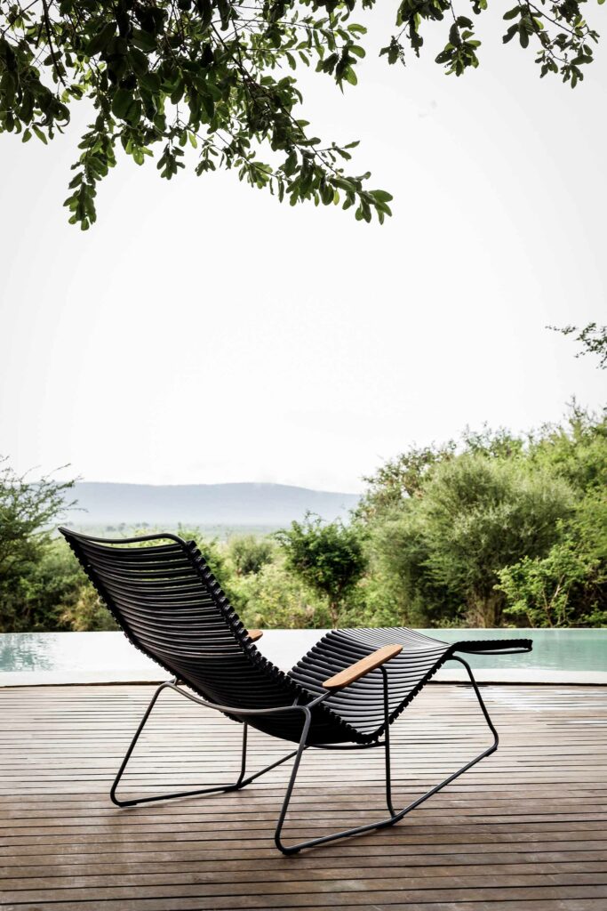Chaise longue Click Sunrocker sur une terrasse en bois au bord d'une piscine à débordement surplombant un paysage naturel.