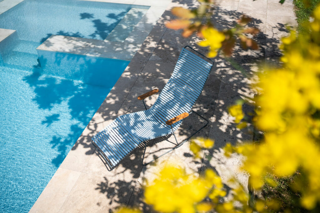 Une chaise longue au bord d'une piscine, partiellement ombragée par des arbres, avec des fleurs jaunes épanouies au premier plan.