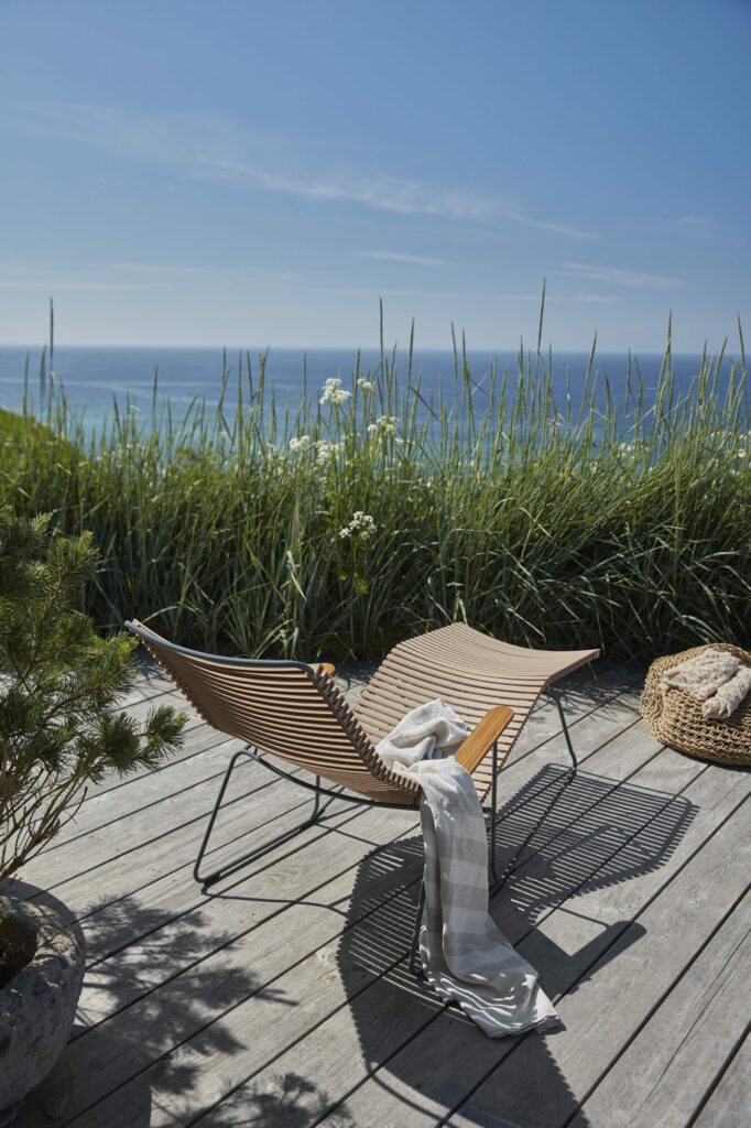 Espace détente extérieur avec chaise longue Click Sunrocker et couverture sur une terrasse en bois avec vue sur la mer, entourée d'herbes hautes.