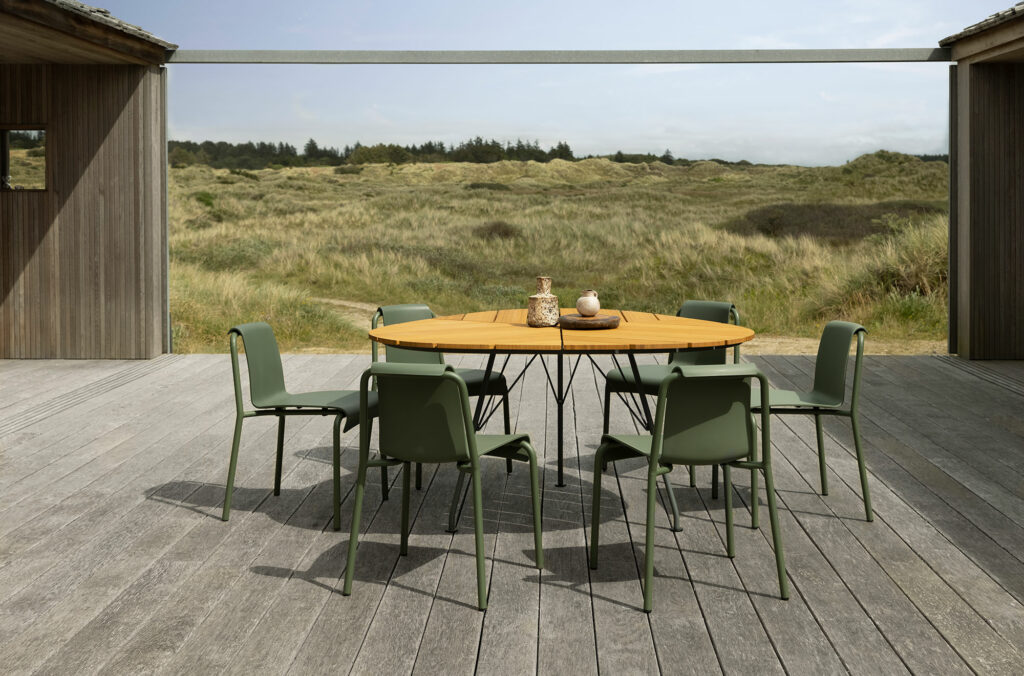 Configuration de salle à manger en plein air avec une table à manger hexagonale LEAF et des chaises sur une terrasse en bois donnant sur un paysage de dunes herbeuses.