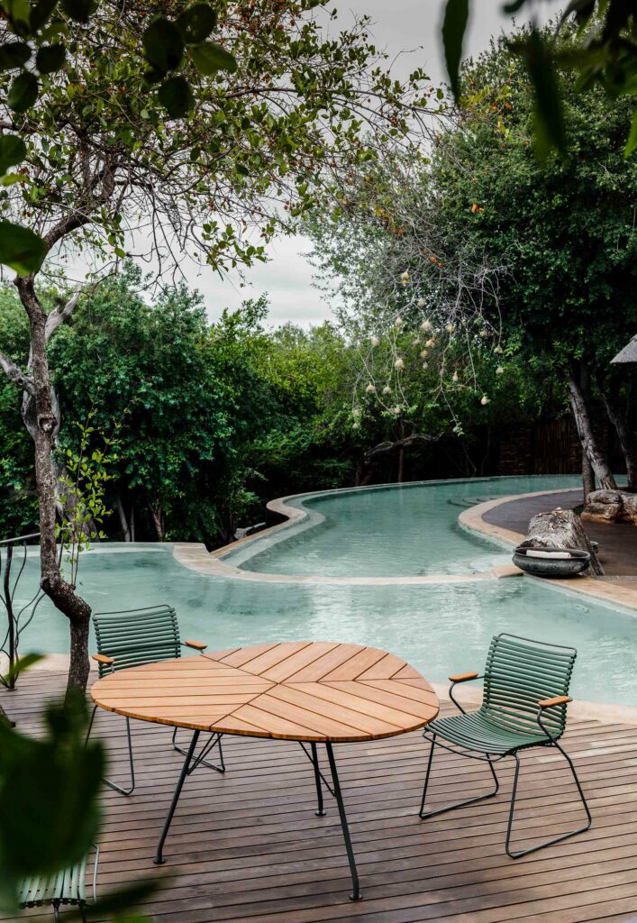Un cadre extérieur serein avec une table et des chaises LEAF au bord d'une piscine sinueuse entourée d'une verdure luxuriante.