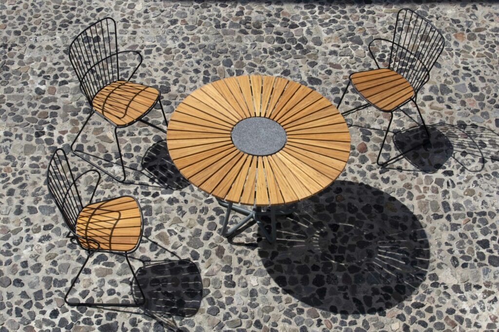 Table à manger circulaire avec chaises en métal sur un trottoir de galets, projetant des ombres au soleil.