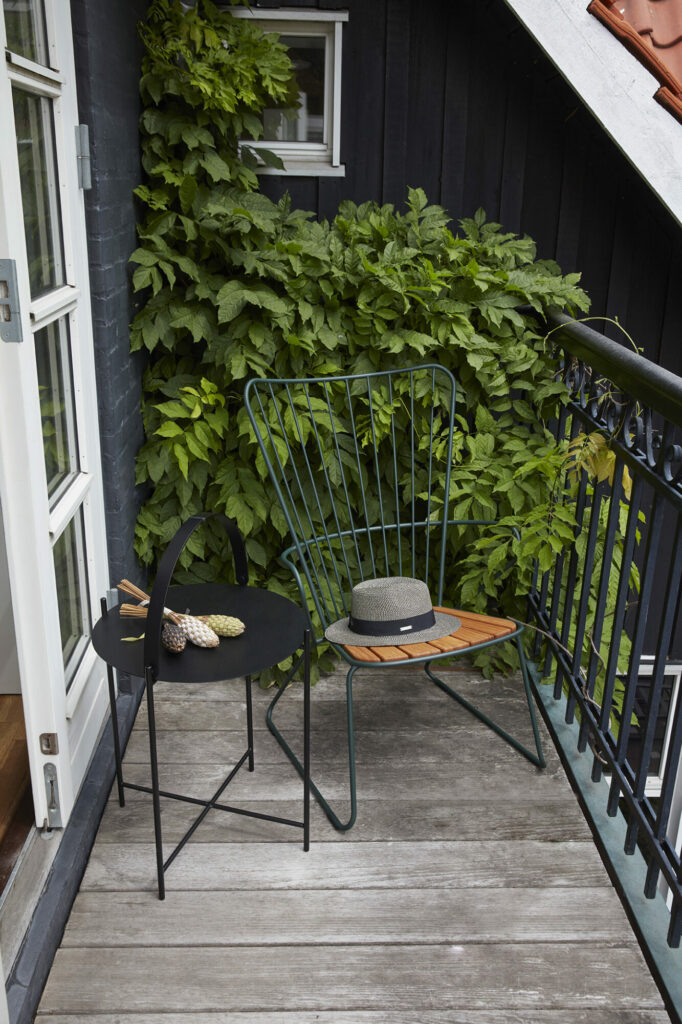 Chaise longue en métal noir Paon avec un chapeau et un livre sur un balcon en bois, entouré d'une verdure luxuriante.
