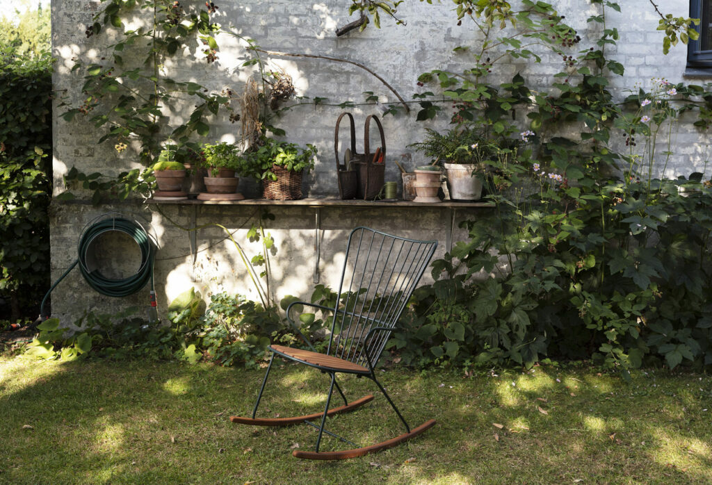 Un coin de jardin tranquille avec une chaise à bascule Paon et une étagère de plantes en pot contre un mur de pierre.