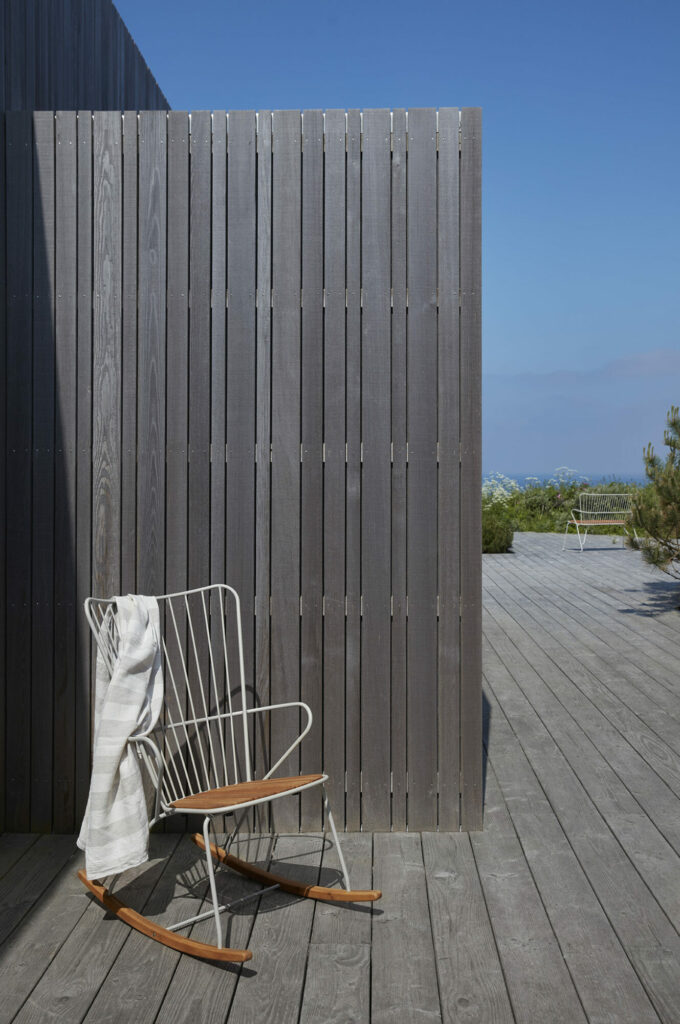 Un coin salon extérieur moderne avec une chaise à bascule Paon et un tissu rayé sur une terrasse en bois, à côté d'un mur vertical en lattes de bois, donnant sur une vue côtière.