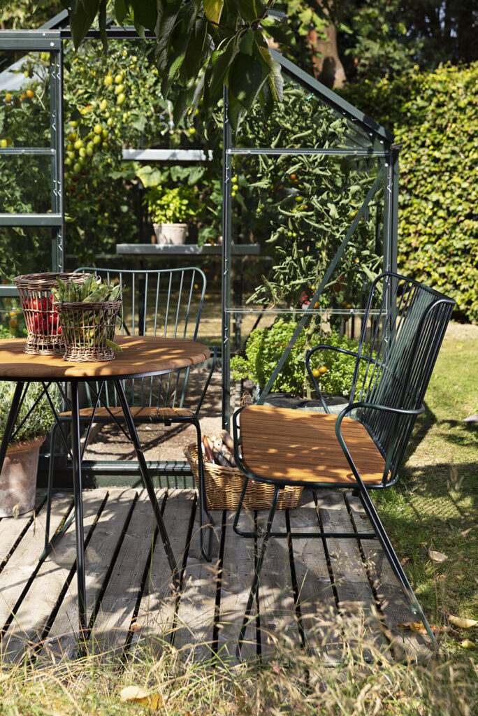 Un petit coin salon extérieur avec une chaise en métal et un banc en bambou blanc à côté d'une serre en verre, dans un jardin luxuriant en toile de fond.