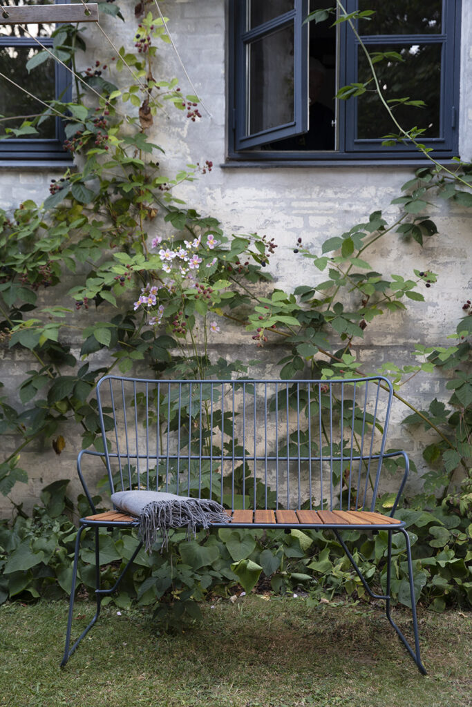 Un banc PAON blanc à structure métallique avec un siège en bambou et une couverture blanche situé devant un mur de pierre avec des plantes grimpantes, près d'une fenêtre avec une personne visible à l'intérieur.