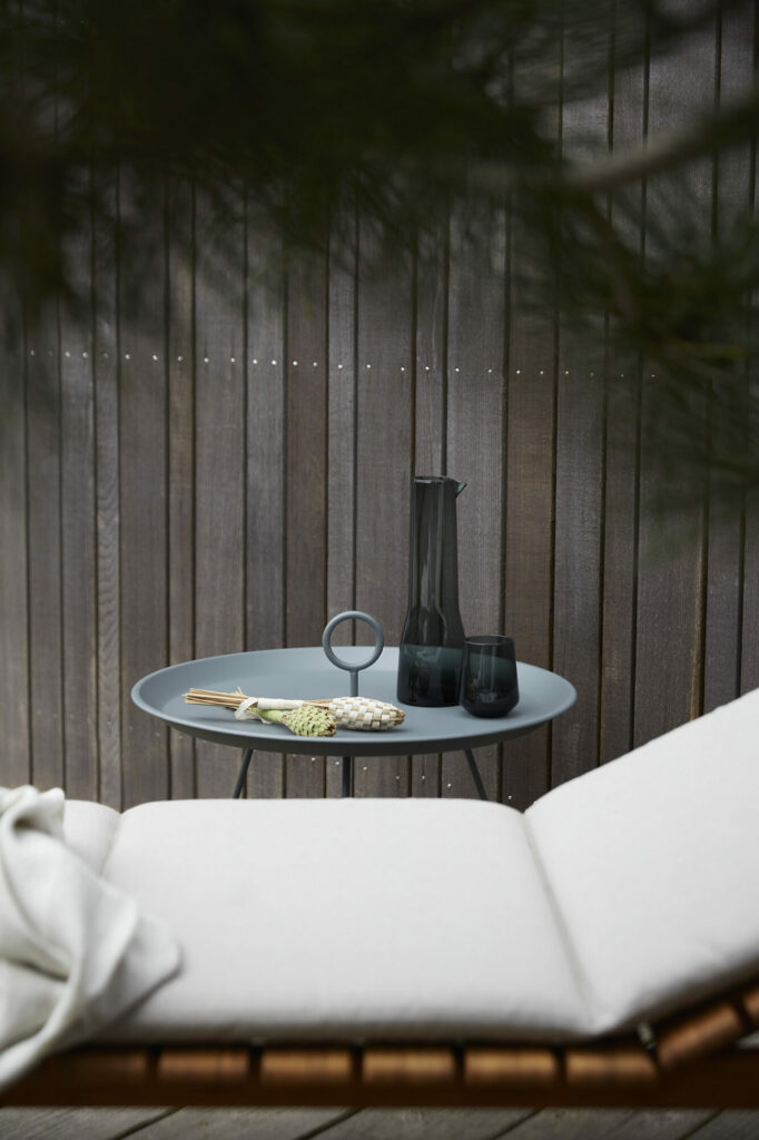 Un coin salon extérieur tranquille comprenant une table EYELET Tray moderne Ø45 avec un vase noir décoratif et un plateau à œillets rempli de bougies, complété par une chaise longue confortable avec un jeté blanc.