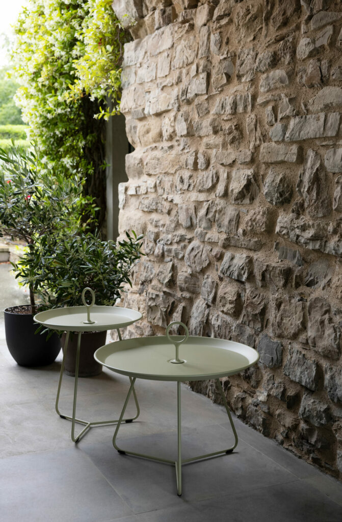 Deux tables EYELET Tray en métal noir, Ø45, avec un verre à vin sur l'une, à côté d'un mur en pierre et d'une plante en pot.