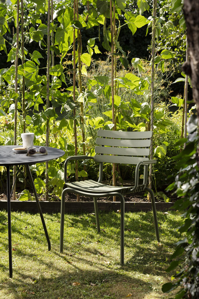 Un jardin tranquille avec une chaise de salle à manger ReCLIPS bleu ciel dotée d'un accoudoir en bambou à côté d'une table avec une tasse à café, entourée de verdure luxuriante et de tiges de haricots.