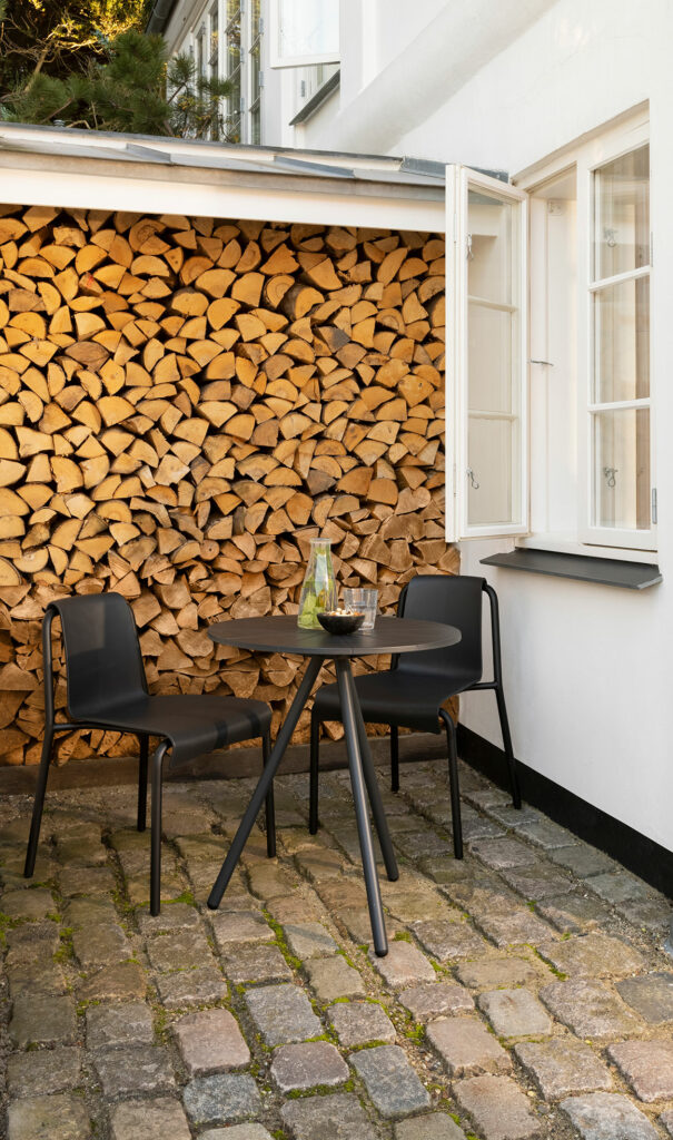 Coin salon extérieur avec chaises et table de salle à manger NAMI noires sur fond de bois de chauffage empilé.