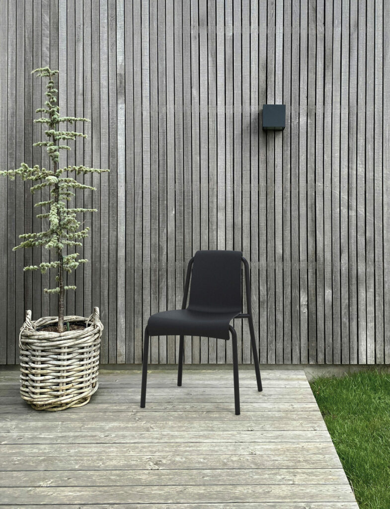 Une chaise de salle à manger NAMI noire et une plante en pot contre un mur à lattes de bois noir avec une lampe extérieure minimaliste installée.