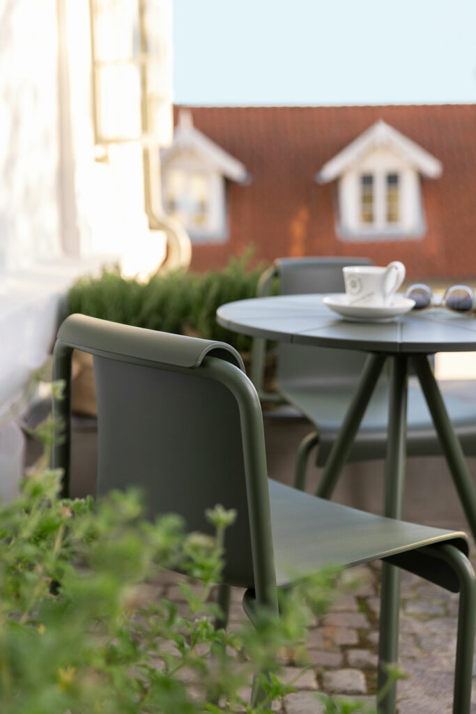 Sièges de café en plein air avec une table vide, une chaise de salle à manger NAMI noire au premier plan, ainsi qu'une tasse à café et des verres sur la table, sur un arrière-plan flou de façades de bâtiments traditionnelles.