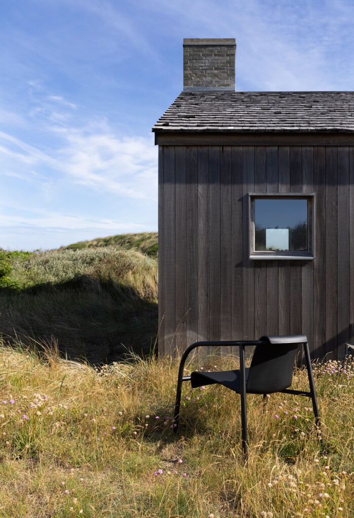 Une cabane en bois avec cheminée se trouve à proximité d'un champ herbeux sous un ciel bleu. Une chaise longue NAMI monochrome en noir épuré est parfaitement positionnée à l'extérieur, face à la cabane.