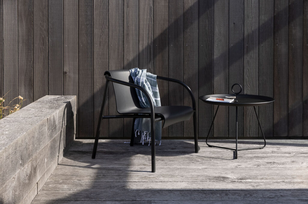 La chaise longue NAMI noire, drapée d'une couverture rayée, se trouve à côté d'une table d'appoint ronde noire remplie de livres sur une terrasse en bois contre un mur en bois.