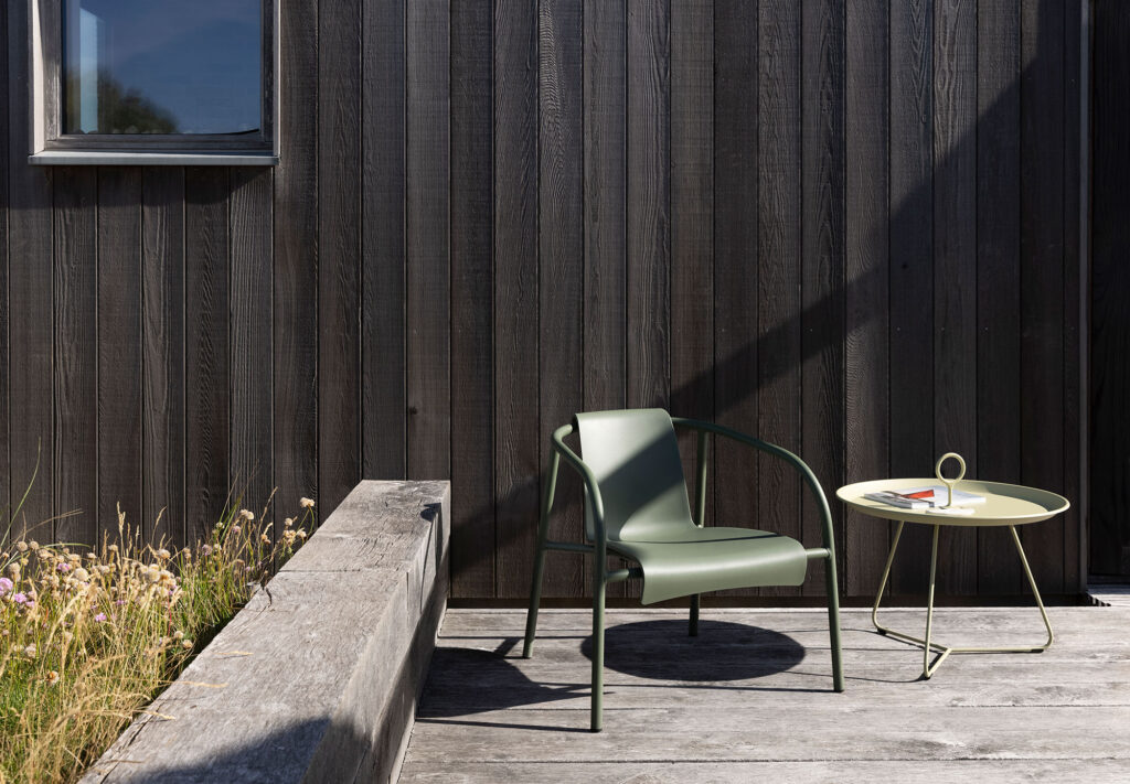 Une chaise longue NAMI noire et une petite table ronde avec un magazine se trouvent sur une terrasse en bois à côté d'un mur noir, accompagnées de jardinières avec de l'herbe et des fleurs.
