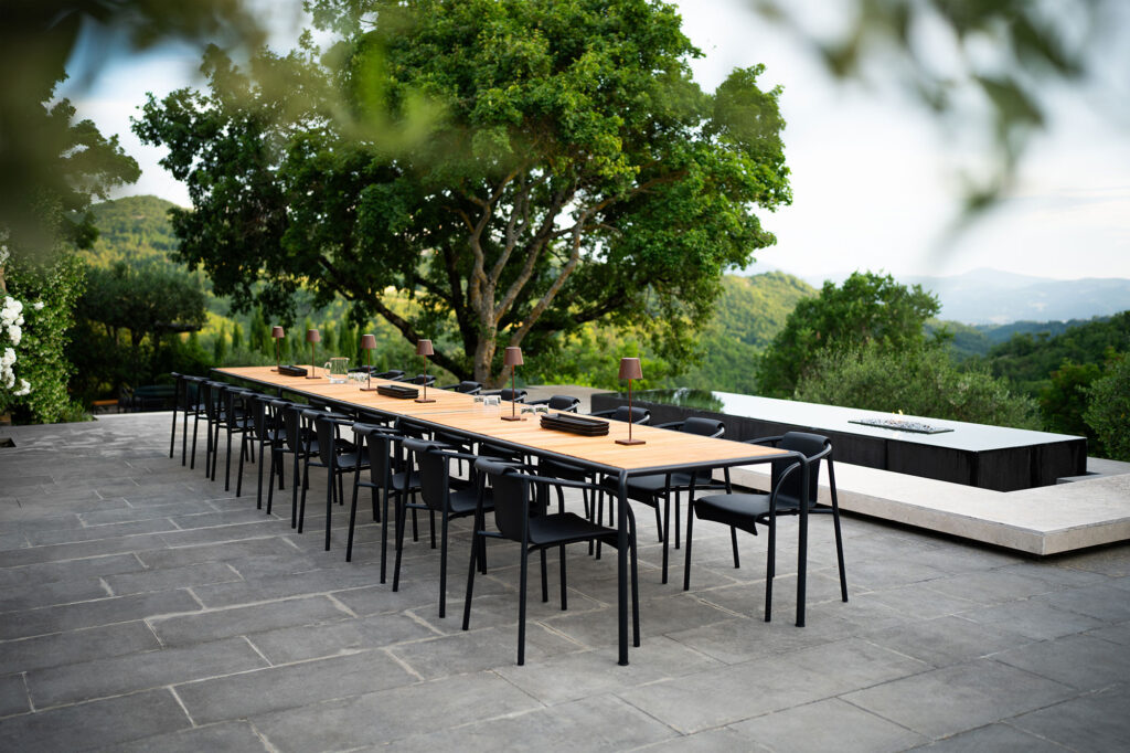 Une longue table à manger d'extérieur avec des chaises de salle à manger NAMI élégantes avec accoudoirs, en noir monochrome, se trouve sur un patio en pierre entouré d'une végétation luxuriante. Un foyer confortable est niché à côté de la table.
