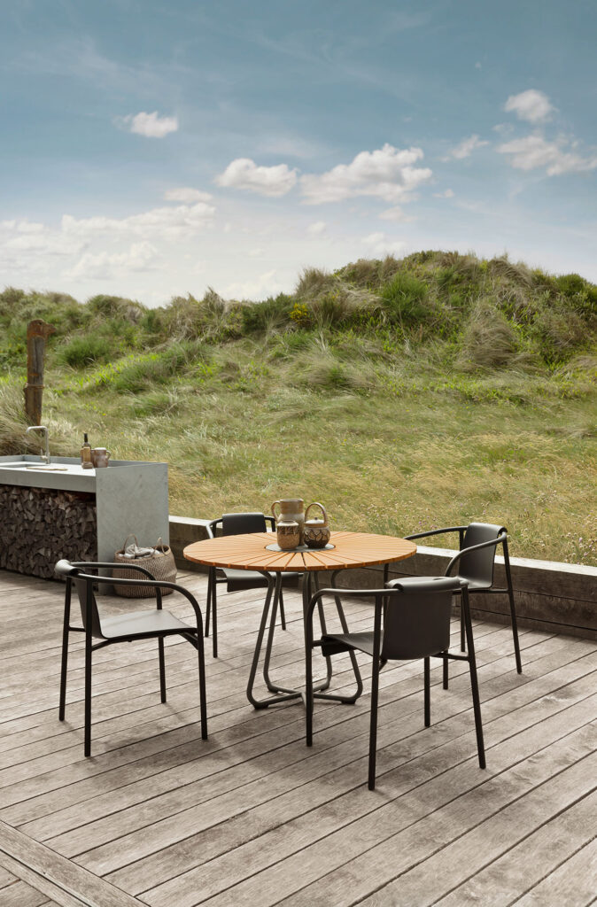 Sur une terrasse en bois, une table ronde est entourée de quatre chaises NAMI noires avec accoudoirs. Le design épuré des chaises contraste joliment avec le paysage herbeux sous un ciel partiellement nuageux.