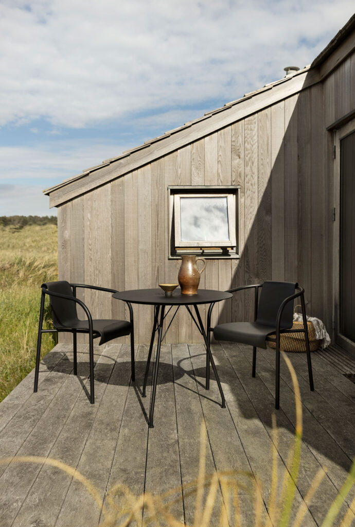 Sur une terrasse en bois à côté d'un bâtiment moderne, un patio extérieur présente deux chaises de salle à manger NAMI noires avec accoudoirs et une table ronde, entourées d'herbe et sur fond de ciel nuageux.