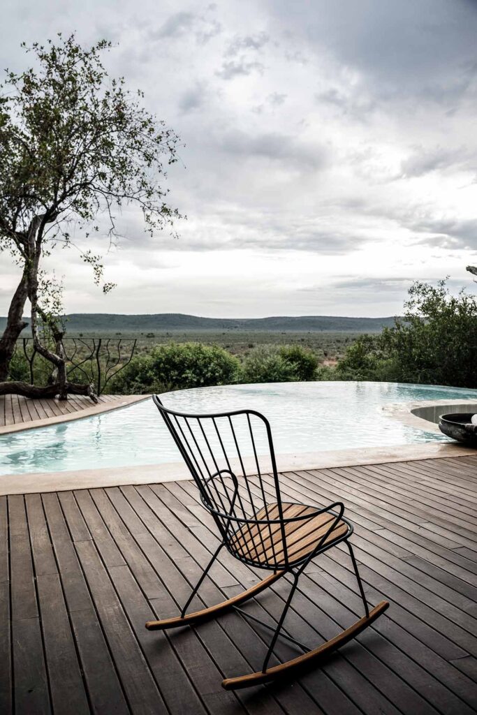 Un seul Rocking Chair Paon orne une terrasse en bois, surplombant une piscine à débordement incurvée avec des collines lointaines sous un ciel nuageux.