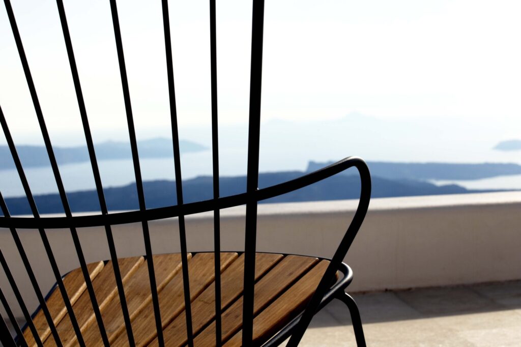 Une chaise à bascule Paon avec un dossier élégant en métal noir est posée sur une terrasse, surplombant un paysage marin serein et des îles lointaines sous un ciel lumineux.