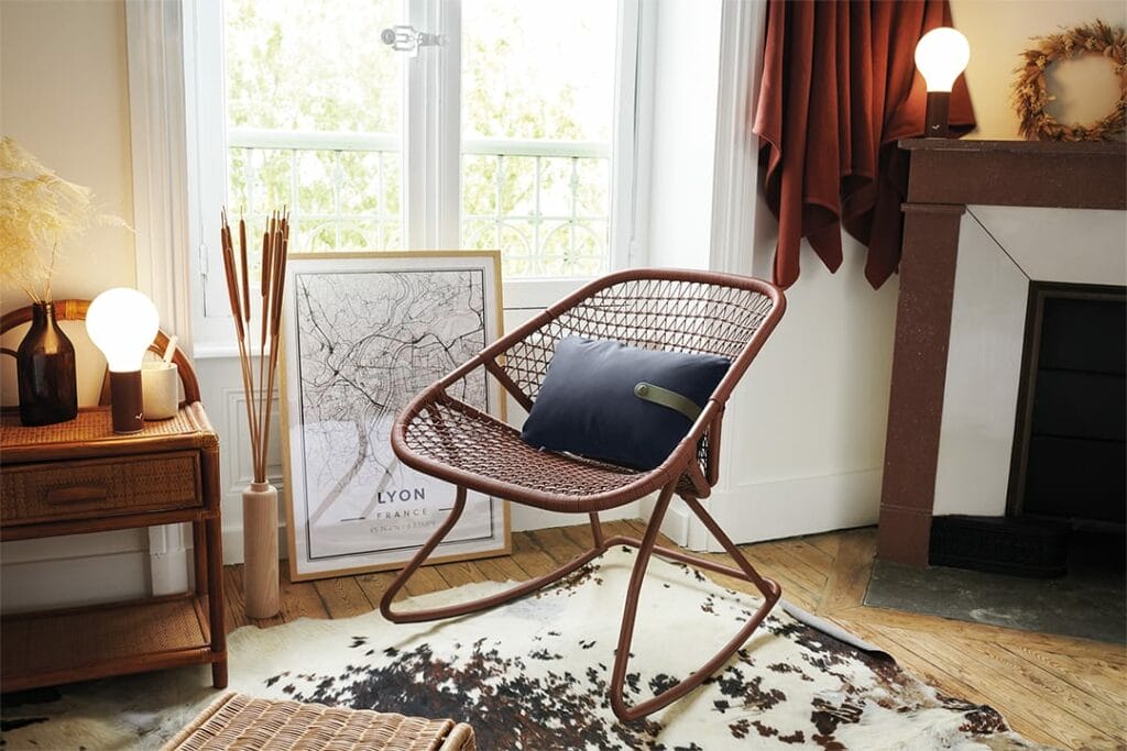 Phrase avec le nom du produit : Une chambre cosy avec un rocking chair élégant des années 60, un coussin bleu, un lampadaire, un plan de Lyon et un tapis en peau de vache à côté d'une cheminée.
