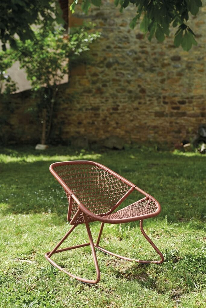 Un fauteuil à bascule filaire de couleur corail des années 60 sur l'herbe devant un mur de pierre, partiellement ombragé par des branches d'arbres en surplomb.