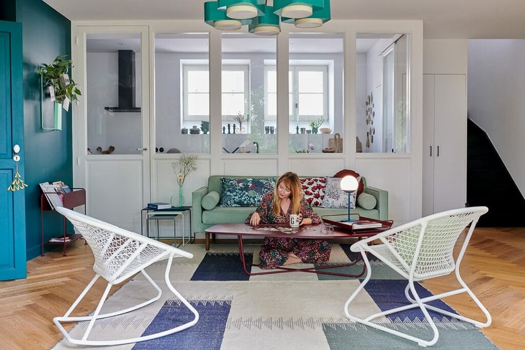 Une femme est assise à une table dans un salon élégant au décor moderne, utilisant un ordinateur portable. La pièce présente des murs bleu sarcelle, des chaises blanches et un fauteuil à bascule vert des années 60.