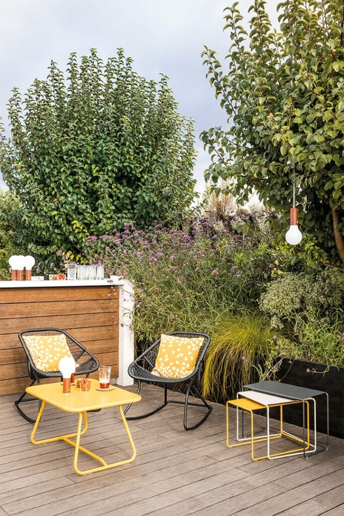 Deux fauteuils à bascule en fil noir des années 60 avec des coussins à motifs jaunes et une table jaune sur une terrasse en bois, entourée d'une verdure luxuriante et d'un bar extérieur.