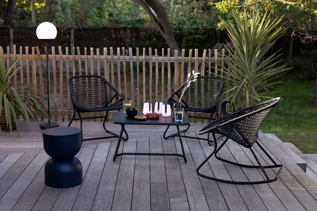 Patio extérieur moderne avec une chaise à bascule noire des années 60 et un ensemble de table, entouré d'une verdure luxuriante et d'une lampe globe blanche inspirée des années 60.