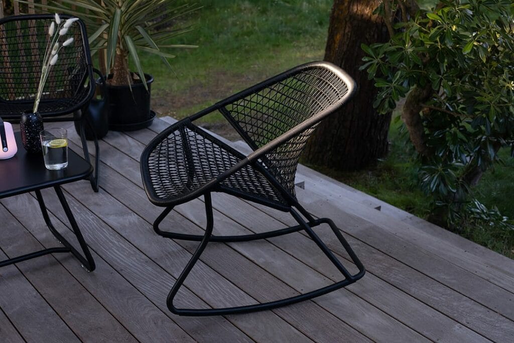 Une chaise à bascule vintage en métal noir des années 60 et une table ronde avec un verre et une bouteille, sur une terrasse en bois entourée de verdure au crépuscule.